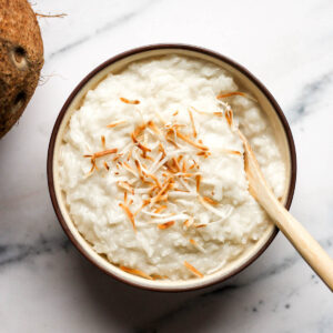 bowl of freshly made coconut rice pudding topped with toasted coconut flakes being stirred with a wooden spoon