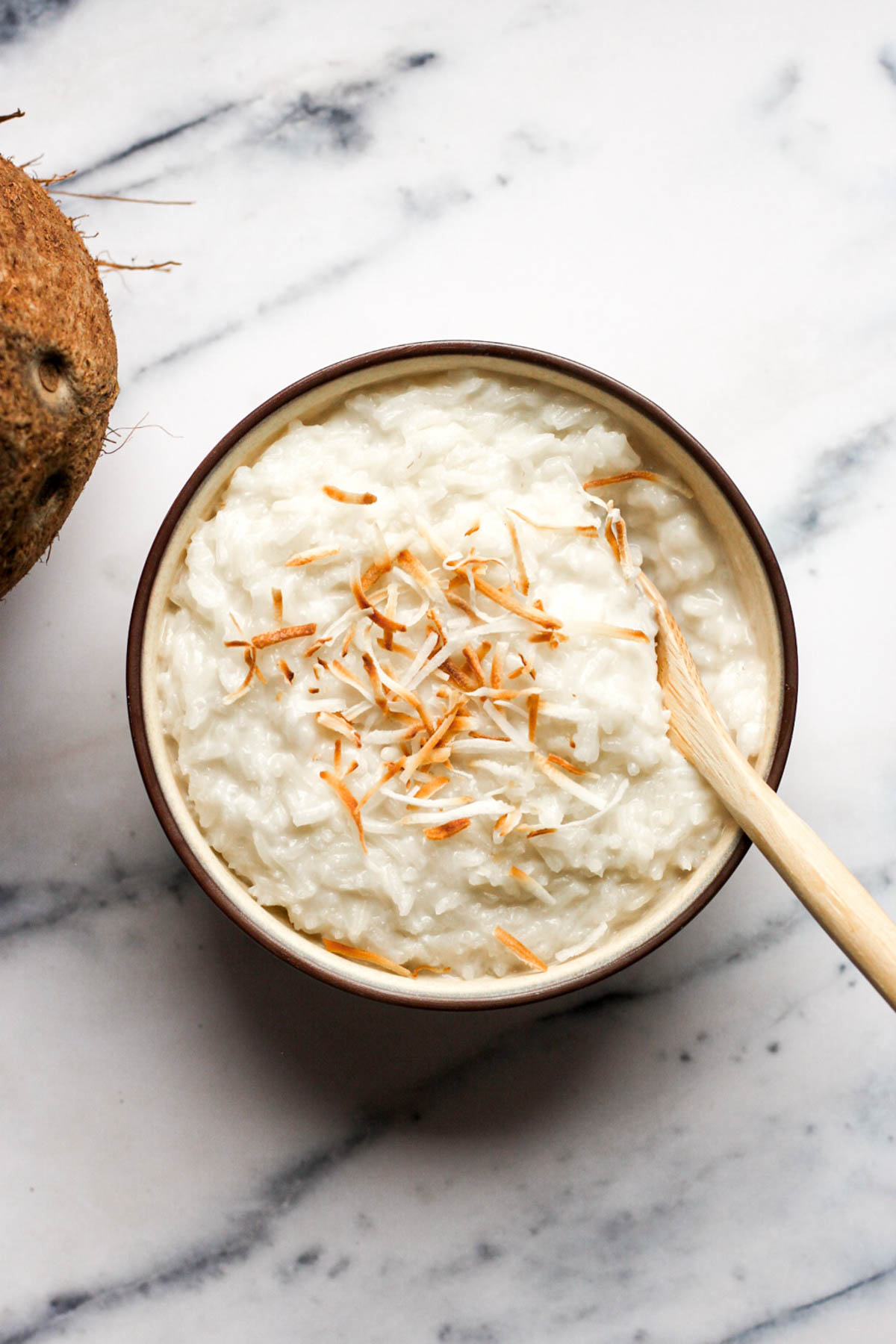 bowl of freshly made coconut rice pudding topped with toasted coconut flakes being stirred with a wooden spoon