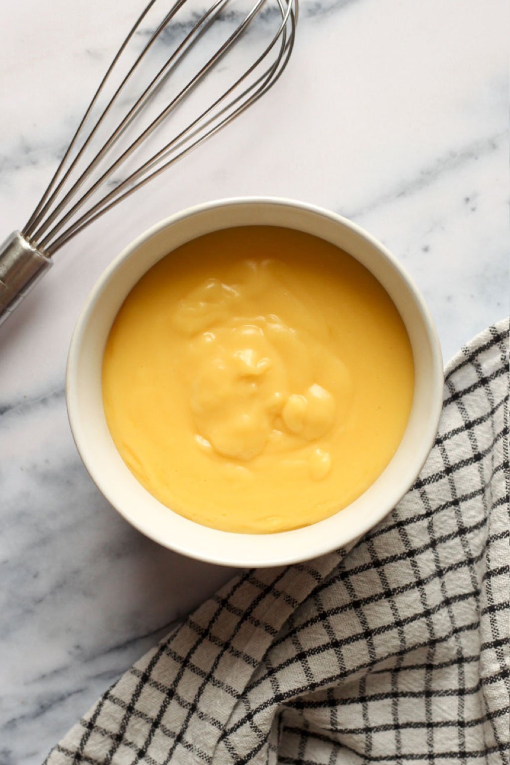 bowl of custard on a marble countertop with a whisk and kitchen towel